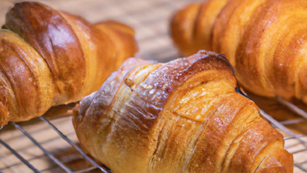Imagem de croissants folheados dourados e crocantes cozidos na Airfryer Lidl. Os croissants têm uma textura escamosa e um interior macio e amanteigado. Eles são servidos em uma cesta de vime com uma xícara de café ao lado.
