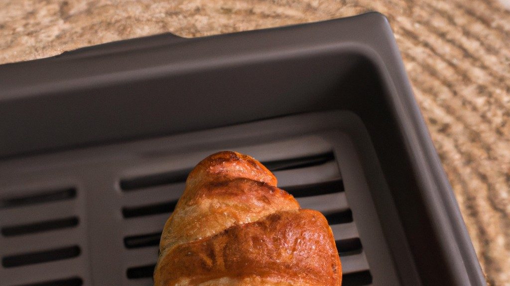 Imagem de croissants quentinhos e dourados preparados na airfryer. Eles são colocados em um prato branco e estão cercados por migalhas de pão. O fundo é uma superfície de madeira clara.