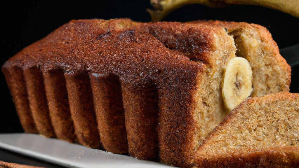 Imagem de um bolo de banana úmido e dourado feito na Airfryer. O bolo é polvilhado com canela e tem uma textura densa e macia. Ele está em um prato branco com uma espátula ao lado.