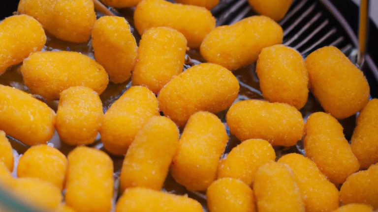 Imagem de salgadinhos de queijo crocantes e dourados preparados na airfryer. Os salgadinhos têm uma forma triangular e são cobertos com queijo derretido e borbulhante. Eles estão dispostos em um prato forrado com papel manteiga.