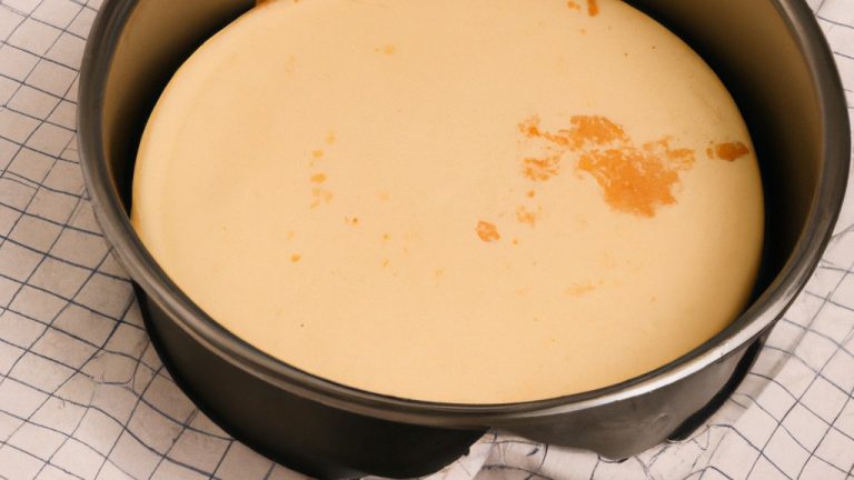 Uma foto aérea de um cheesecake assado em uma airfryer. O cheesecake tem uma crosta dourada e um topo cremoso e levemente dourado. É servido em uma assadeira pequena.