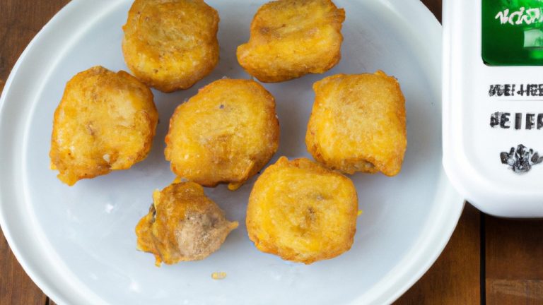 Imagem de nuggets de frango congelados dourados e crocantes servidos em uma tigela branca com um lado de molho de tomate. Os nuggets são empanados e têm um interior branco e macio. A tigela está sobre uma superfície de madeira com um pano de prato xadrez vermelho e branco ao lado.