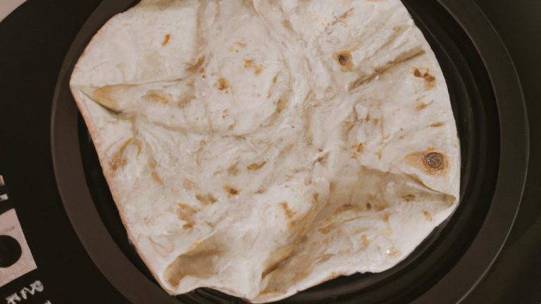 Imagem de um pão folha dourado cozido na Airfryer, uma fritadeira sem óleo. O pão tem um formato redondo e está ligeiramente achatado, com uma superfície texturizada e bolhas de ar visíveis. Ele está disposto sobre uma superfície branca com um fundo desfocado.